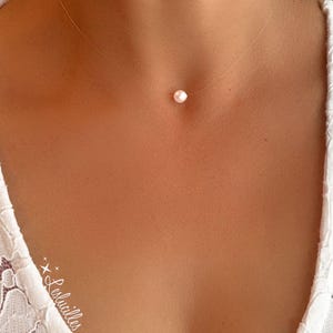 Puede incluir: Un collar simple y delicado con un solo pequeño colgante de perla blanca que cuelga de una cadena transparente casi invisible.