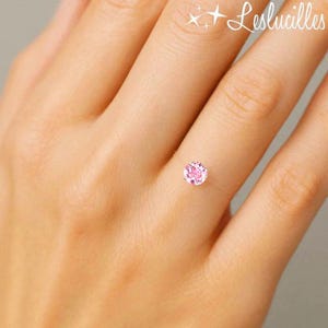 以下が含まれることがあります： 単一の、丸い、薄いピンク色の宝石を特徴とする繊細なリング。 宝石はほぼ見えないバンドにセットされており、浮遊効果を生み出しています。 リングは人の指に表示されています。 テキスト「Leslucilles」は右上隅にあります。