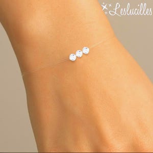 May include: A delicate bracelet featuring three clear, round, sparkling gemstones strung on a nearly invisible cord. The bracelet is shown on a person's wrist, with the brand name "Leslucilles" in the upper right corner.