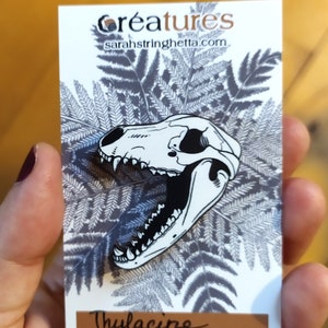 Peut inclure: Une épingle en émail noir et blanc représentant un crâne stylisé de tigre de Tasmanie, également connu sous le nom de thylacine. L'épingle est sur une carte blanche avec un motif de fougère et le texte "creatures sarahstringhetta.com" et "Thylacine".