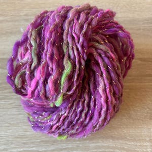 May include: A skein of yarn with a purple, pink, and green color scheme. The yarn is interwoven with gold metallic thread.