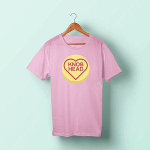 May include: Pink t-shirt featuring a yellow heart-shaped sweet design that reads "Knob Head" in red lettering.