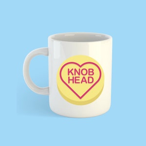 May include: White ceramic mug with a yellow heart-shaped candy design. The candy has a pink outline and the text "KNOB HEAD" in pink letters.