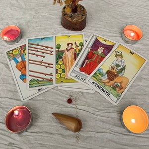 May include: A tarot card reading setup featuring several tarot cards with illustrations and titles, a wooden pendulum, and lit candles. The cards depict various figures and symbols, with the word "STRENGTH" visible on one card.