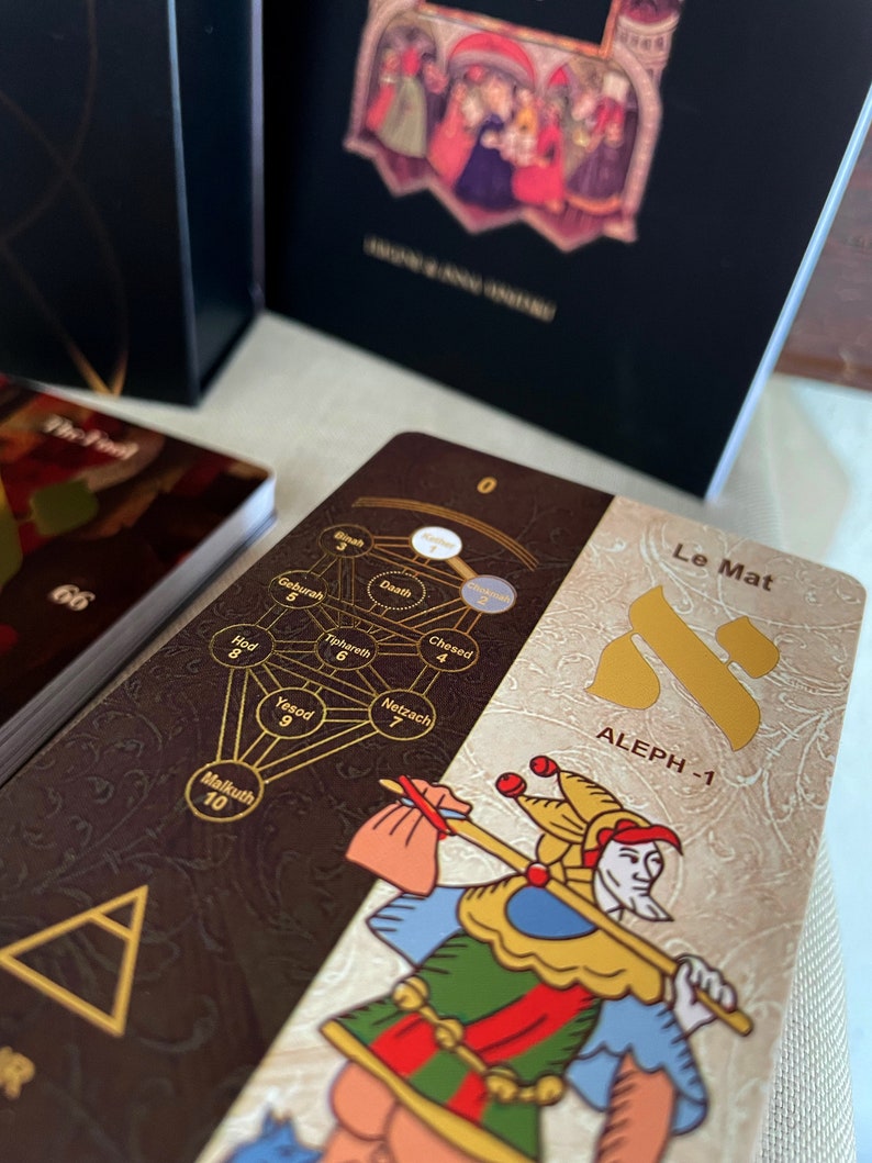 May include: A tarot card with a brown background and gold accents. The card features a jester holding a staff and a tree of life with Hebrew letters and numbers. The card is titled "Le Mat" and "ALEPH -1".