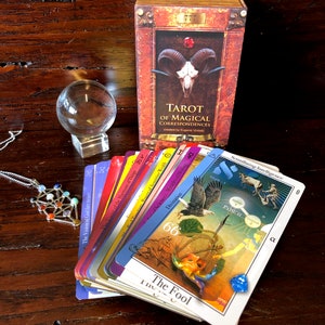 May include: A tarot deck with a red, white, and blue color scheme. The cards are fanned out to show the different suits and numbers. The box for the deck is red and gold with a skull and horns on the front. The text on the box reads "Tarot of Magical Correspondences" and "created by Eugene Viniski".