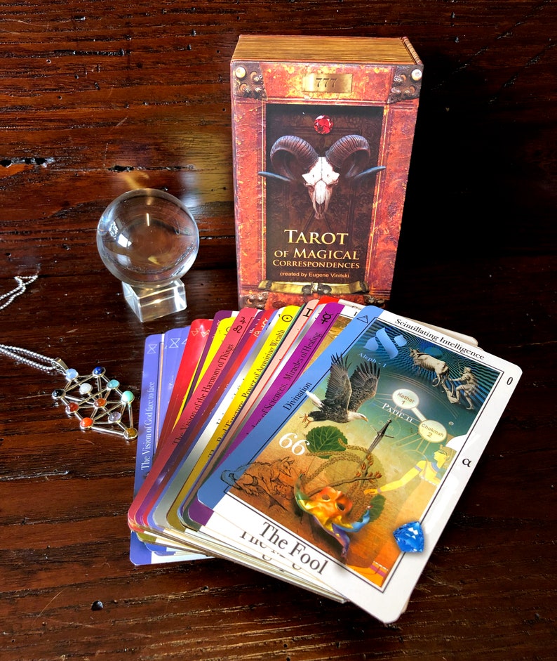 May include: A tarot deck with a red, white, and blue color scheme. The cards are fanned out to show the different suits and numbers. The box for the deck is red and gold with a skull and horns on the front. The text on the box reads "Tarot of Magical Correspondences" and "created by Eugene Viniski".