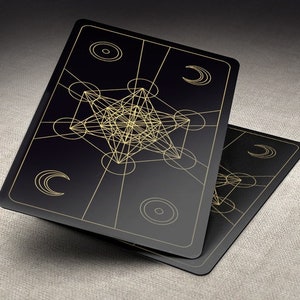 May include: Two black playing cards with gold geometric designs. The cards feature a complex, intricate pattern of overlapping circles and lines, creating a visually striking effect.