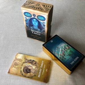 May include: Three tarot card decks with gold accents. The top deck is titled "Money Tarot" by Eugene Vinitski. The bottom left deck is titled "Money Tarot" by Eugene Vinitski. The bottom right deck has a blue background with a card image and the text "The Fool".