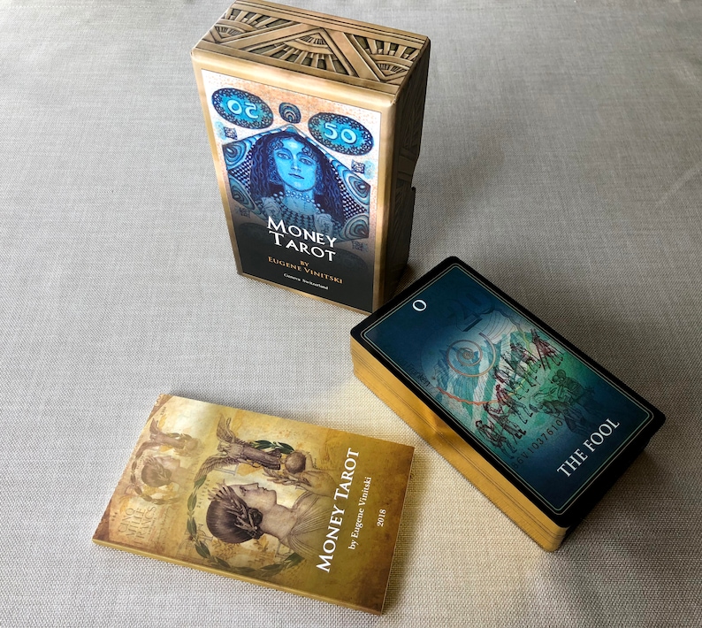 May include: Three tarot card decks with gold accents. The top deck is titled "Money Tarot" by Eugene Vinitski. The bottom left deck is titled "Money Tarot" by Eugene Vinitski. The bottom right deck has a blue background with a card image and the text "The Fool".