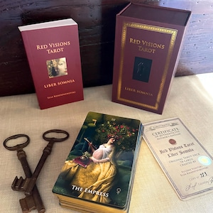 May include: A Red Visions Tarot Liber Somnia deck set including a maroon guidebook, a matching storage box with gold trim, a deck of cards featuring The Empress, two antique-style metal keys, and a certificate of authenticity numbered 333.
