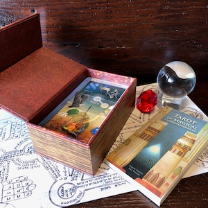 May include: A wooden box with a deck of tarot cards inside. The box is open and the cards are visible. The cards are illustrated with images of animals, plants, and other symbols. The box is sitting on a white surface with a book and a crystal ball.