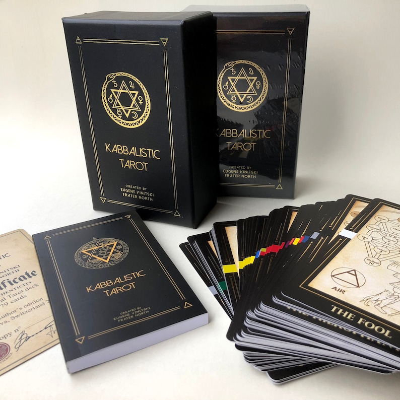 May include: A black box with gold lettering that reads "Kabbalistic Tarot" created by Eugene Vintski Frater North. The box is next to a deck of tarot cards with a black and white design. The top card is "The Fool" with a black and white illustration of a person walking with a staff.