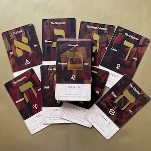 May include: A collection of tarot cards featuring Hebrew letters and illustrations. The cards are arranged in a circular pattern on a brown surface. The cards are in various colors, including red, gold, and black.
