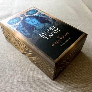 May include: A gold box with a black label that reads "Money Tarot" by Eugene Vinitksi, Geneva Switzerland. The box features a blue and gold illustration of a woman with a crown.