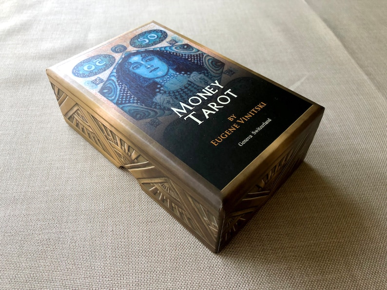 May include: A gold box with a black label that reads "Money Tarot" by Eugene Vinitksi, Geneva Switzerland. The box features a blue and gold illustration of a woman with a crown.