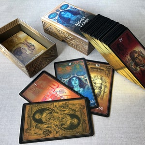 May include: A gold box with the words "Money Tarot" on the front, with a deck of tarot cards in a black and gold box. The cards have images of people and symbols, and the box has a gold design.
