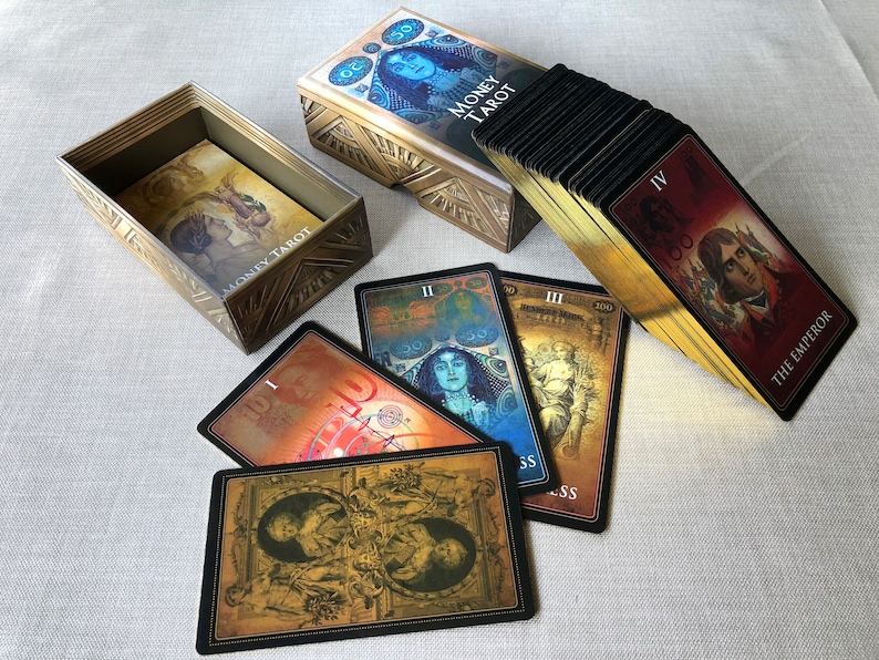 May include: A gold box with the words "Money Tarot" on the front, with a deck of tarot cards in a black and gold box. The cards have images of people and symbols, and the box has a gold design.