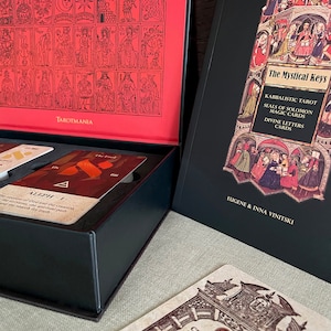 May include: A black box with a red lid containing a deck of The Mystical Keys Kabbalistic Tarot cards. The box is open and the cards are visible. The lid of the box has a black and white illustration of the 22 Major Arcana cards. The book, "The Mystical Keys Kabbalistic Tarot Seals of Solomon Magic Cards Divine Letters Cards" by Eugene & Inna Vinitski is visible in the background.
