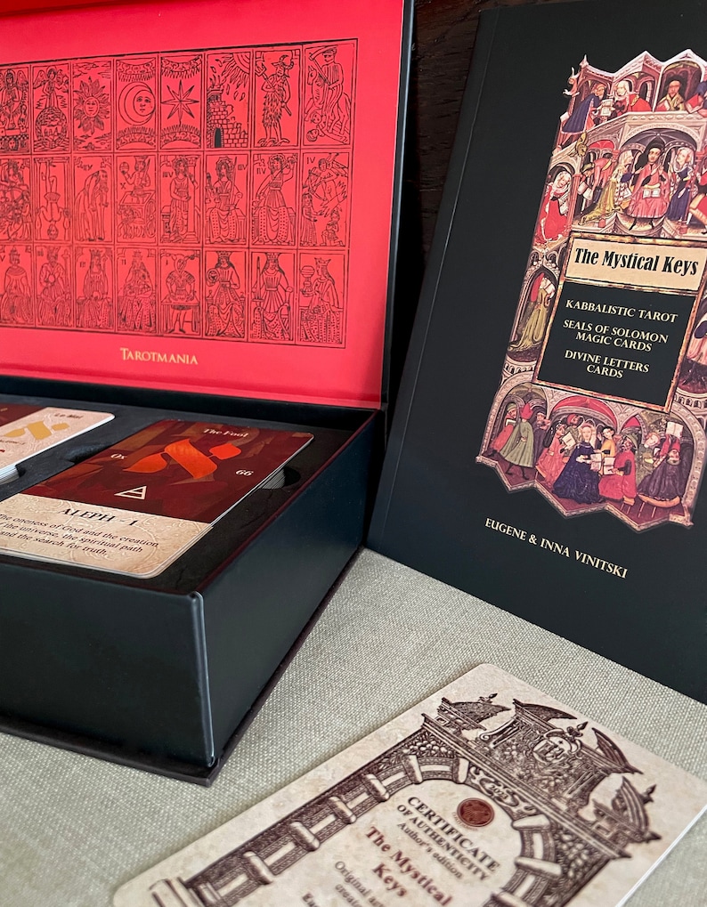 May include: A black box with a red lid containing a deck of The Mystical Keys Kabbalistic Tarot cards. The box is open and the cards are visible. The lid of the box has a black and white illustration of the 22 Major Arcana cards. The book, "The Mystical Keys Kabbalistic Tarot Seals of Solomon Magic Cards Divine Letters Cards" by Eugene & Inna Vinitski is visible in the background.