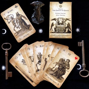 May include: An oracle deck titled "The Dance of Death" with illustrations of skeletons and figures. The deck includes cards with zodiac symbols, a key, and a decorative metal object. The cards are arranged on a black surface.