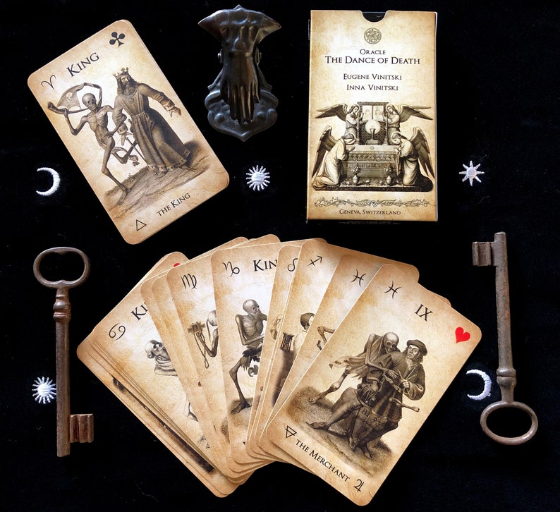 May include: An oracle deck titled "The Dance of Death" with illustrations of skeletons and figures. The deck includes cards with zodiac symbols, a key, and a decorative metal object. The cards are arranged on a black surface.