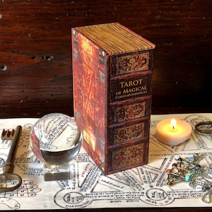 May include: A closed book with a brown leather cover and gold accents. The title of the book is "Tarot of Magical Correspondences". The book is sitting on a table with a crystal ball, a candle, and a key.