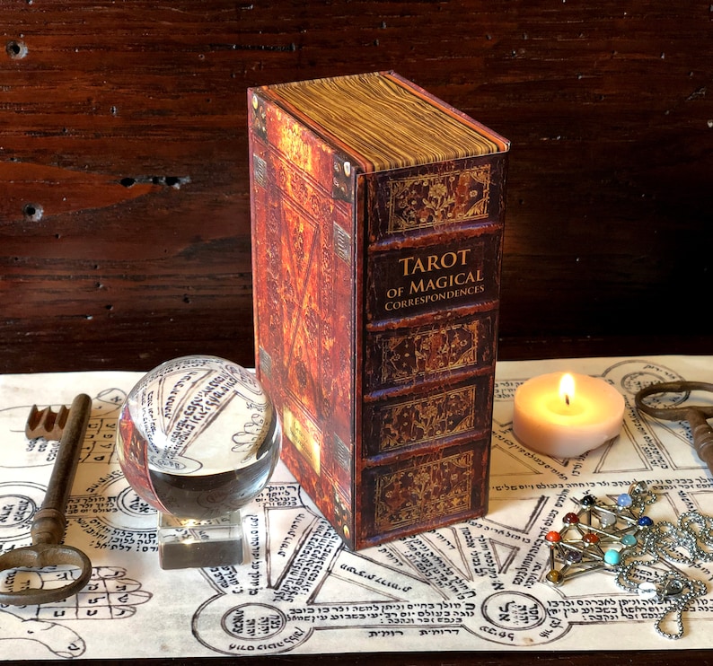 May include: A closed book with a brown leather cover and gold accents. The title of the book is "Tarot of Magical Correspondences". The book is sitting on a table with a crystal ball, a candle, and a key.