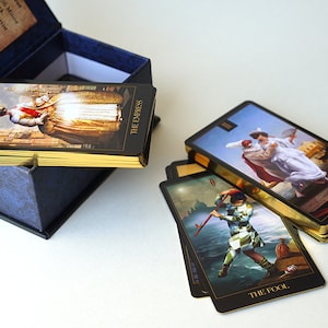 May include: A set of tarot cards with gold gilded edges in a blue box. The cards feature illustrations of people and scenes. The cards are arranged in a stack with the top card showing "The Empress" and the bottom card showing "The Fool".