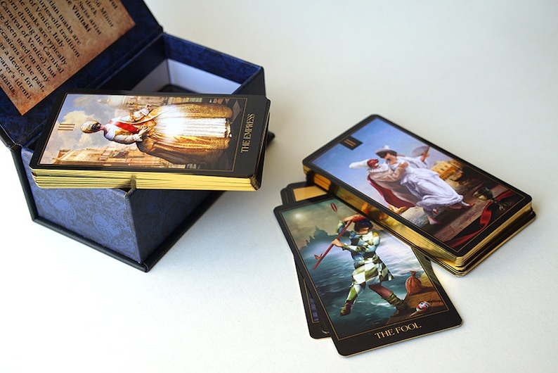 May include: A set of tarot cards with gold gilded edges in a blue box. The cards feature illustrations of people and scenes. The cards are arranged in a stack with the top card showing "The Empress" and the bottom card showing "The Fool".
