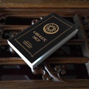 May include: A black book with gold lettering titled "Kabbalistic Tarot" by Eugene Vinitski, Frater North. The book features a gold star of David with a snake wrapped around it.