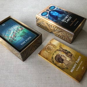 May include: Two gold boxes with intricate designs containing decks of Money Tarot cards. One box is open and shows a card with the Fool depicted on it. The other box is closed and has the title "Money Tarot" on the front. The third box is closed and has the title "Money Tarot" on the front.