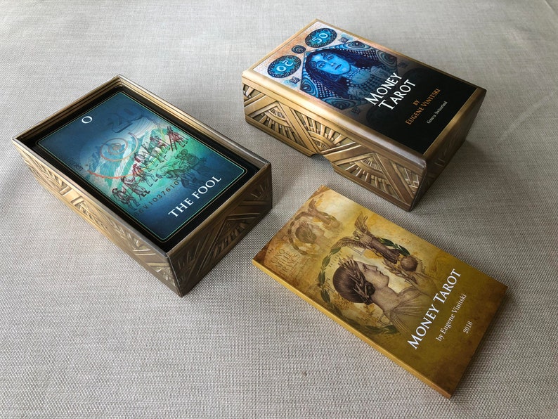May include: Two gold boxes with intricate designs containing decks of Money Tarot cards. One box is open and shows a card with the Fool depicted on it. The other box is closed and has the title "Money Tarot" on the front. The third box is closed and has the title "Money Tarot" on the front.
