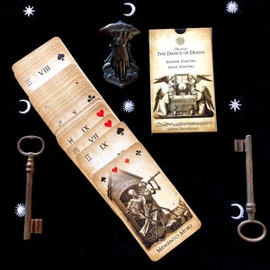 May include: An oracle card deck titled "The Dance of Death" with illustrations of skeletons and angels. The deck is fanned out, revealing card suits and Roman numerals. Two antique keys and a decorative hand-shaped object are also present.