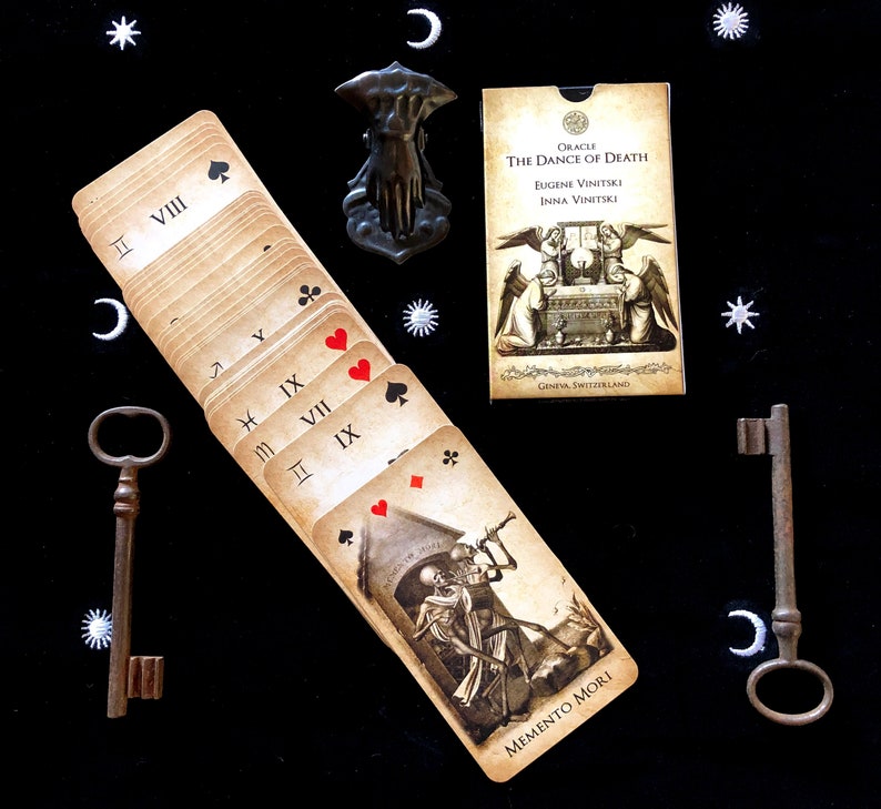 May include: An oracle card deck titled "The Dance of Death" with illustrations of skeletons and angels. The deck is fanned out, revealing card suits and Roman numerals. Two antique keys and a decorative hand-shaped object are also present.