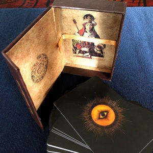 May include: A brown box with a gold ribbon and a card inside. The card features a painting of a person wearing a crown and holding a book. The box is open and a stack of black cards with a gold eye design are visible.