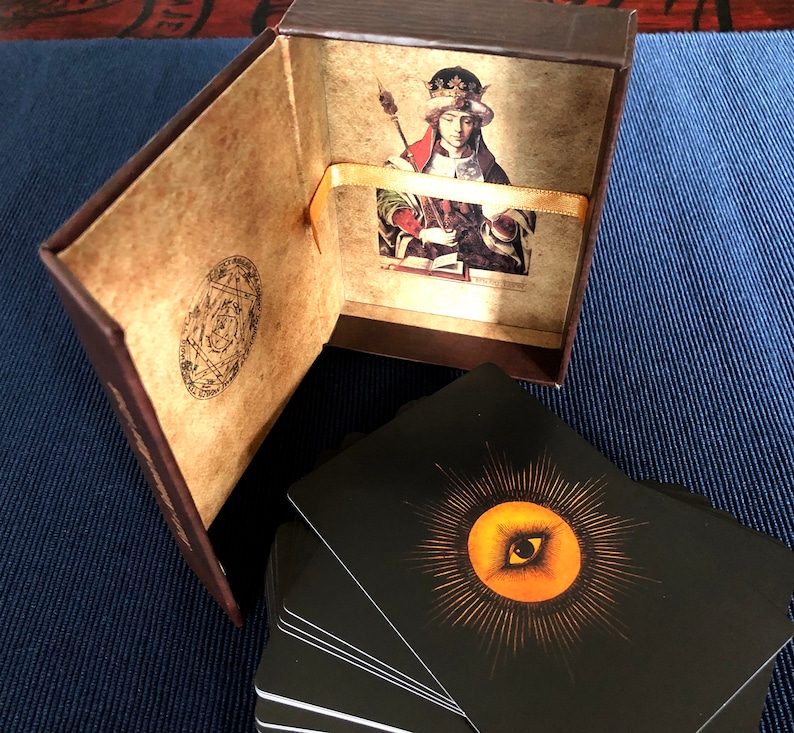 May include: A brown box with a gold ribbon and a card inside. The card features a painting of a person wearing a crown and holding a book. The box is open and a stack of black cards with a gold eye design are visible.