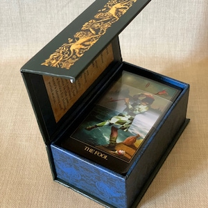 May include: A closed box with a blue and gold design. The box is open to reveal a tarot card with a colorful illustration of a figure in a jester's outfit. The card is labeled "THE FOOL".
