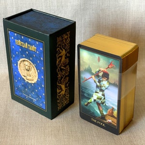 May include: A green box with gold accents and a blue lid with gold stars and the text "Venetian Tarot" on the front. The box is next to a deck of tarot cards with gold edges and a black card with the text "The Fool" on the front.