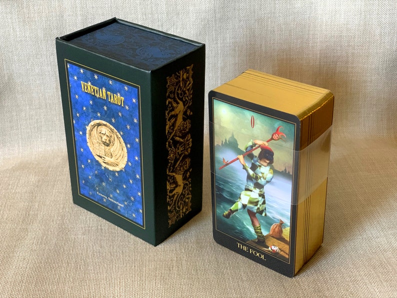 May include: A green box with gold accents and a blue lid with gold stars and the text "Venetian Tarot" on the front. The box is next to a deck of tarot cards with gold edges and a black card with the text "The Fool" on the front.