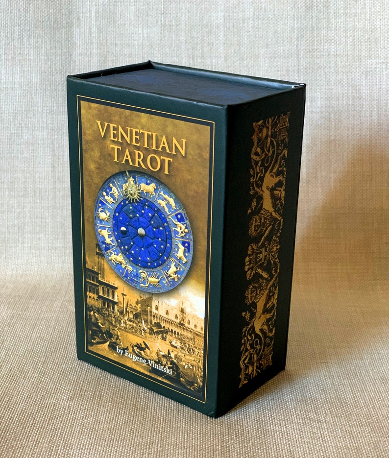 May include: A black box with gold accents and the text "Venetian Tarot" on the front. The box features a blue and gold illustration of a zodiac wheel with a cityscape in the background. The text "by Eugene Vinitski" is at the bottom of the box.