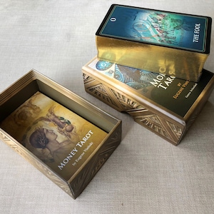 May include: Three tarot card decks in their boxes. The top deck is a blue and gold deck with the text "The Fool" on the box. The middle deck is a black and gold deck with the text "Money Tarot" on the box. The bottom deck is a gold box with the text "Moono Tarot" on the box.