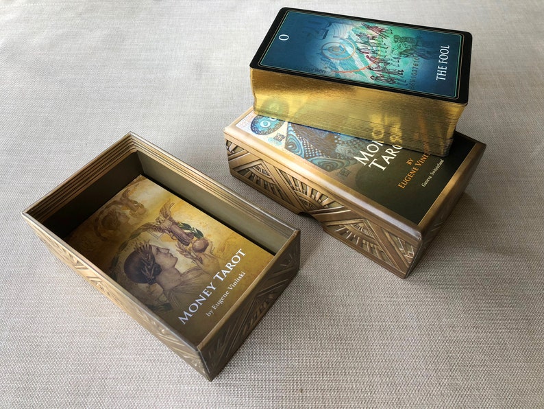 May include: Three tarot card decks in their boxes. The top deck is a blue and gold deck with the text "The Fool" on the box. The middle deck is a black and gold deck with the text "Money Tarot" on the box. The bottom deck is a gold box with the text "Moono Tarot" on the box.