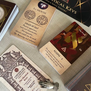 May include: A set of tarot cards with a certificate of authenticity. The cards feature a gold and red design with the text "The Mystical Keys" and "Aleph - 1".