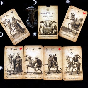 May include: An oracle card deck titled "The Dance of Death" with illustrations of skeletons and figures. The deck includes cards with the text "King," "The Emperor," "The Jester," "The Executioner," and "The Merchant." The cards are arranged on a black surface with decorative keys.