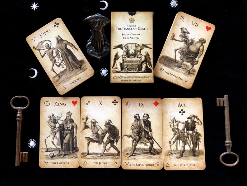 May include: An oracle card deck titled "The Dance of Death" with illustrations of skeletons and figures. The deck includes cards with the text "King," "The Emperor," "The Jester," "The Executioner," and "The Merchant." The cards are arranged on a black surface with decorative keys.