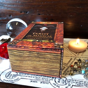 May include: A closed box of Tarot of Magical Correspondences cards, created by Eugene Vinitaki. The box is brown with gold accents and has a textured surface. The title of the deck is printed on the front of the box in gold lettering.