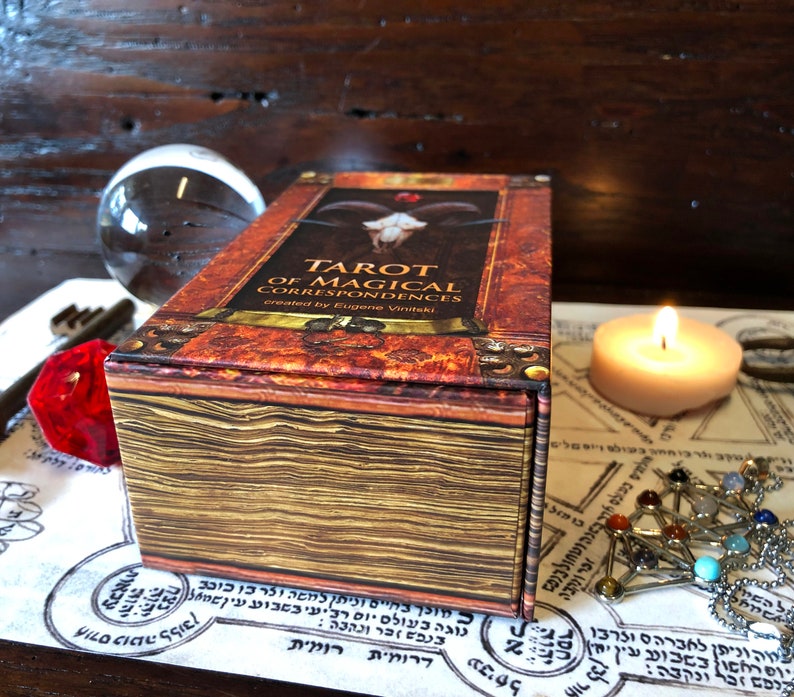 May include: A closed box of Tarot of Magical Correspondences cards, created by Eugene Vinitaki. The box is brown with gold accents and has a textured surface. The title of the deck is printed on the front of the box in gold lettering.
