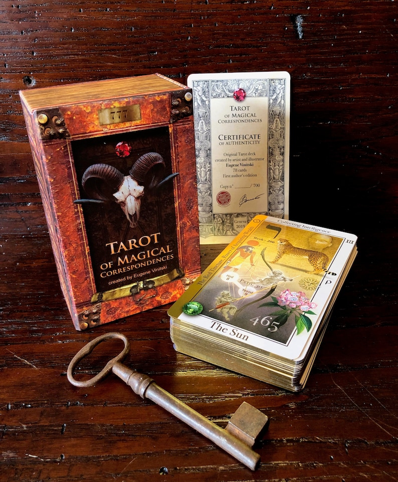 May include: A tarot deck box with a gold and red design featuring a skull with horns. The box is labeled "Tarot of Magical Correspondences" and "created by Eugene Vinitski". The box is sitting on a wooden surface with a stack of tarot cards and a key.