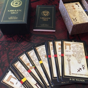 May include: A spread of black and gold tarot cards with Hebrew letters and illustrations of trees. The cards are laid out on a dark red wooden surface. The cards are from the Kabbalistic Tarot deck.